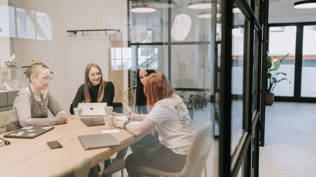 ier Frauen sitzen an einem Holztisch in einem modernen Büro mit Glaswand und arbeiten gemeinsam an Laptops.