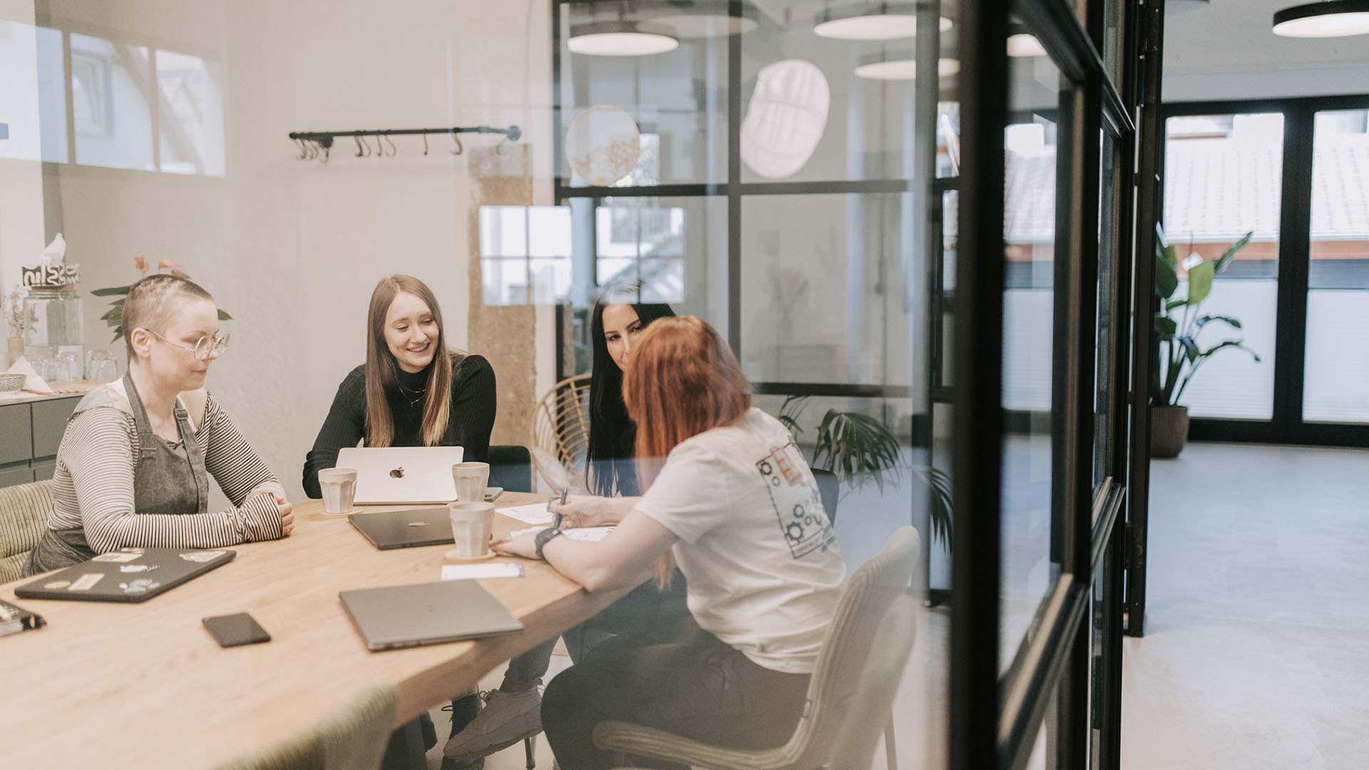 ier Frauen sitzen an einem Holztisch in einem modernen Büro mit Glaswand und arbeiten gemeinsam an Laptops.