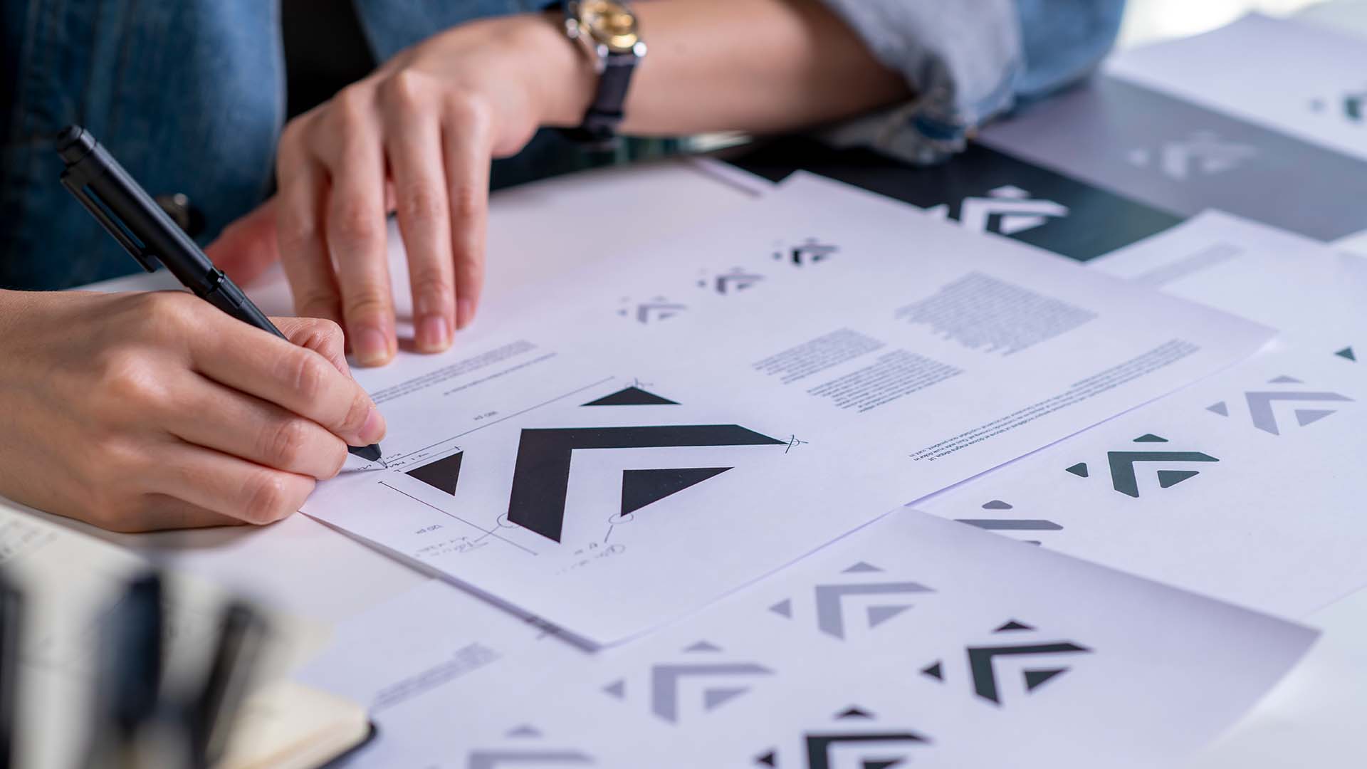 Nahaufnahme einer Hand mit Armbanduhr, die mit einem Bleistift eine Logo-Skizze auf Papier zeichnet, umgeben von ausgedruckten schwarzen Logo-Varianten.